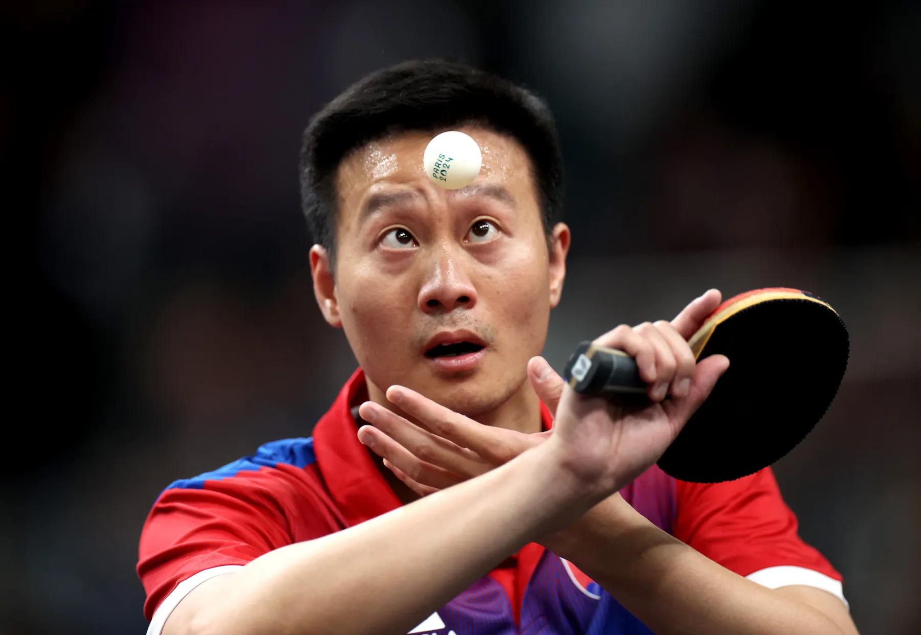 Table tennis player focusing intently on a spinning ball above his forehead, showcasing concentration and skill during a competitive match. He is wearing a red and blue sports uniform, holding a paddle in one hand while positioning his other hand for the next move.