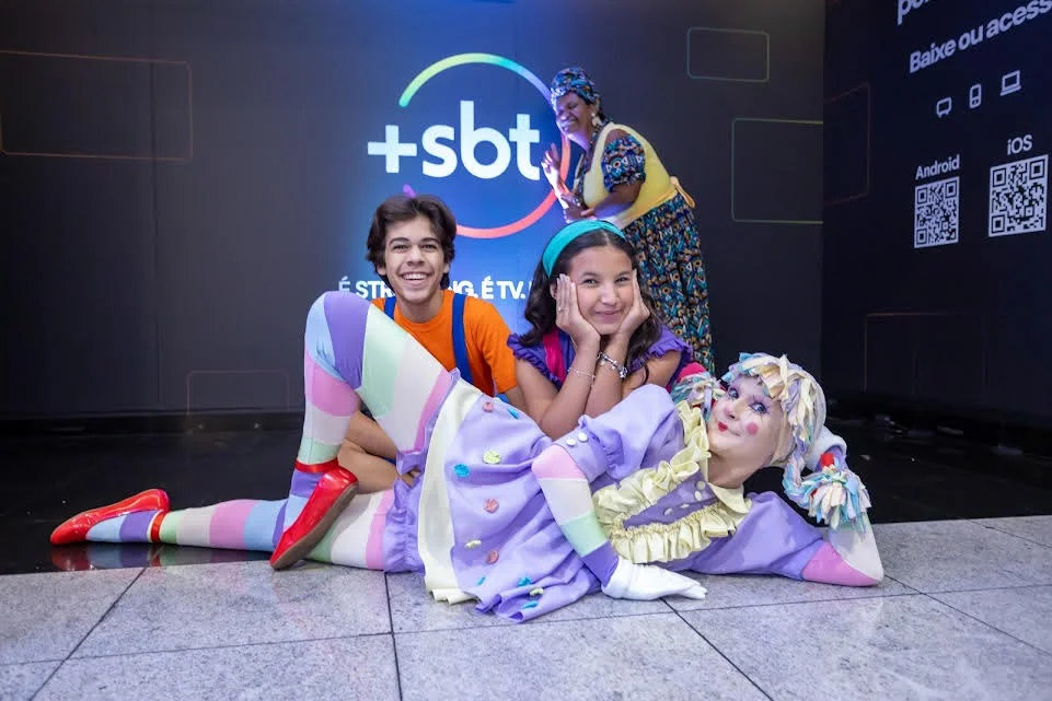 Children and a clown pose playfully in front of an SBT promotional backdrop, showcasing a vibrant and festive atmosphere. The boy wears an orange shirt, while the girl sports a pink top, both smiling joyfully. The clown, dressed in colorful attire, lies on the floor, adding to the fun and entertainment theme.