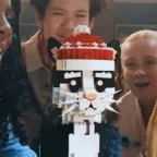 Crianças admirando gato de LEGO com touca vermelha.