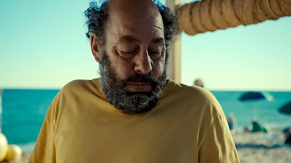 Homem de barba na praia, usando camiseta amarela.