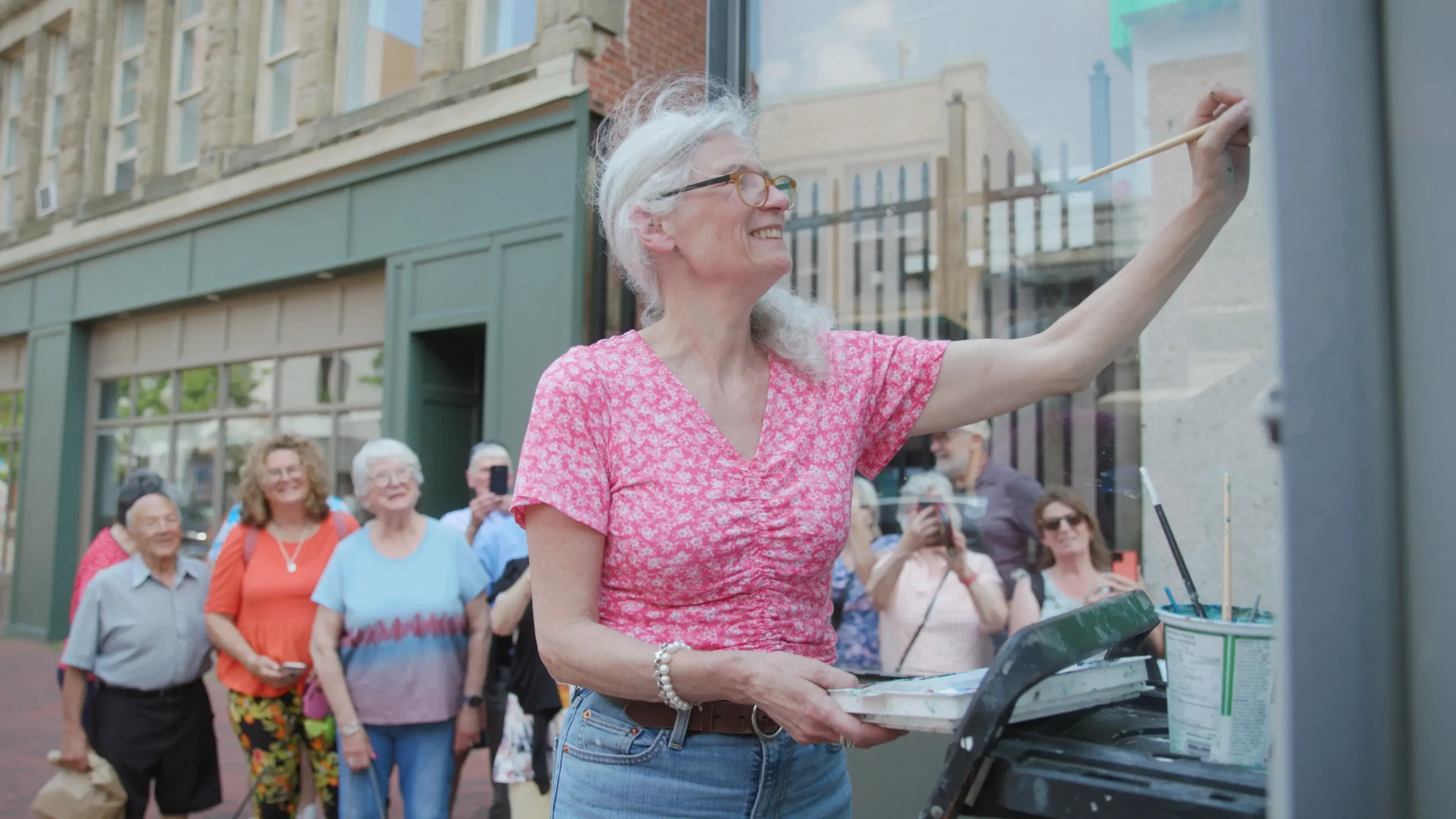 Mulher pintando vitrine com público sorridente.