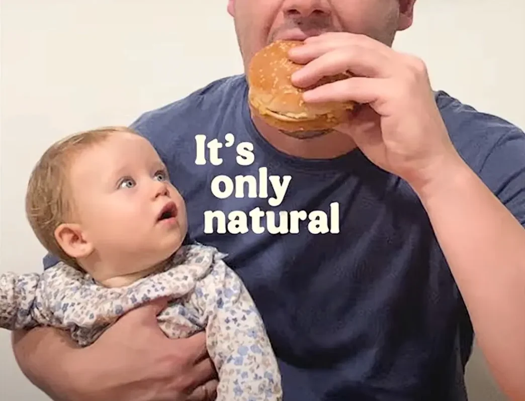 Bebê observa adulto comendo hambúrguer.
