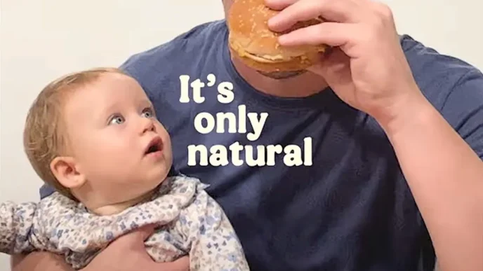 Bebê observa adulto comendo hambúrguer.