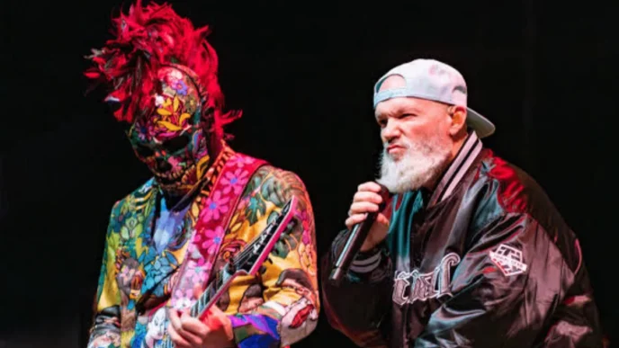 Músicos performando em show, um com roupa colorida.