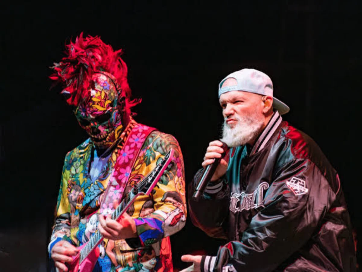 Músicos performando em show, um com roupa colorida.