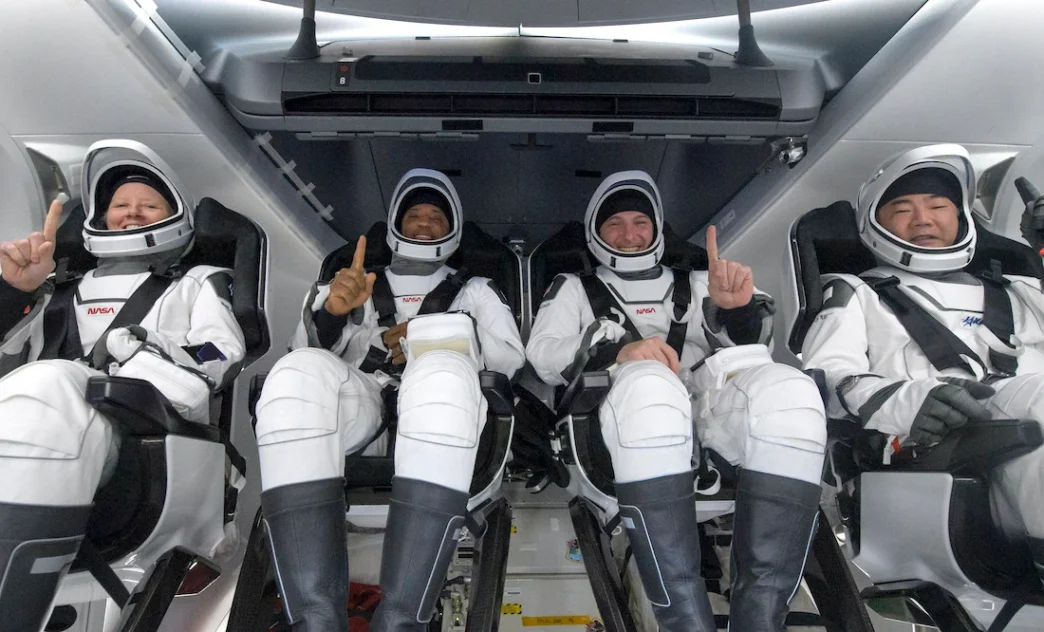 Quatro astronautas em trajes espaciais sorrindo na nave.