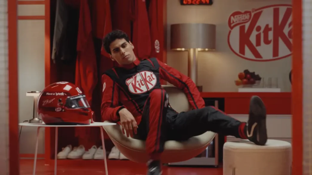 Piloto relaxa em vestiário KitKat, capacete ao lado.