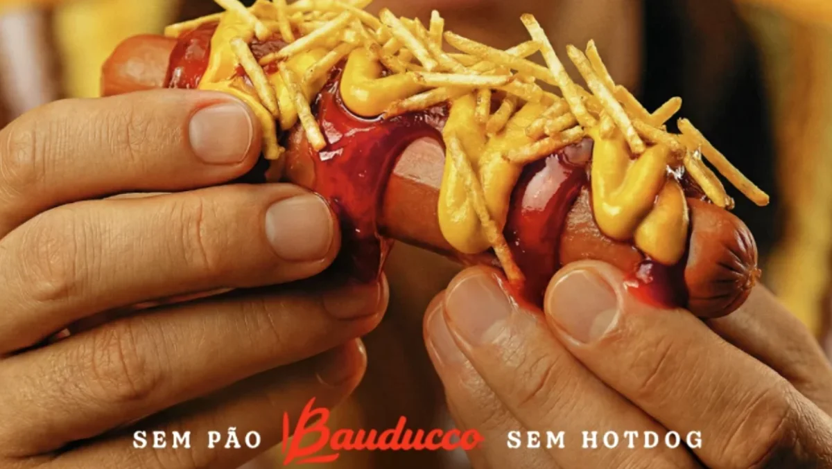 Mãos segurando salsicha com molhos e batata palha.