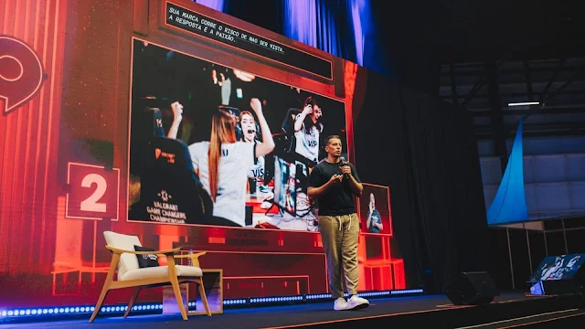 Palestrante em palco com telão e plateia ao fundo
