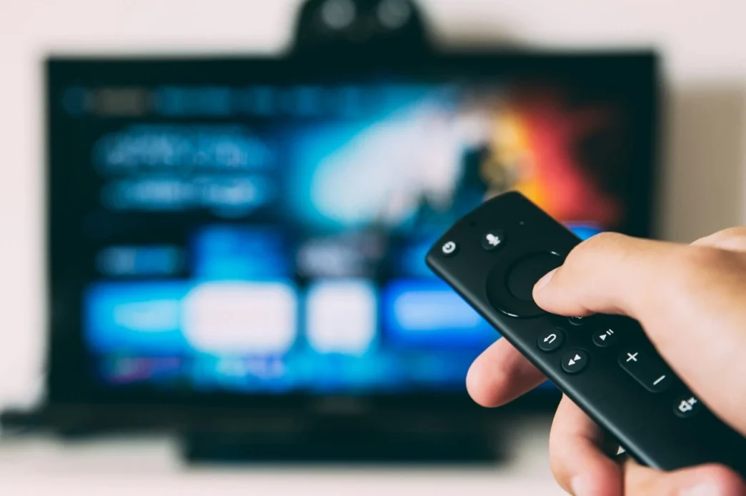 Pessoa segura controle remoto diante de televisão ligada, representando o consumo de documentários esportivos em streaming.